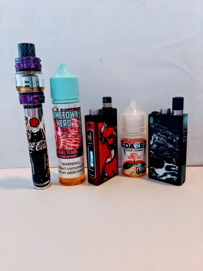 An array of vaping devices sit alongside vaping juice flavors apple and watermelon berry. These flavors attract teenagers because of their fruity appeal.

