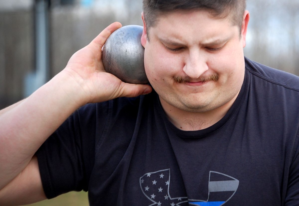 Track coach Thomas Fleming loads a shot put onto his shoulder getting ready to throw for a demonstration at practice in the NBC Sports Complex. Thomas holds the eight pound spherical shot put only holding on with his palm. 
