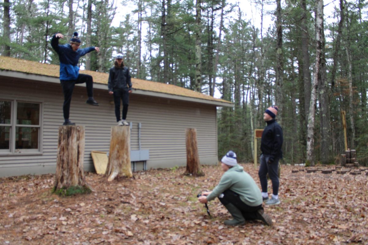 Junior Mike Fiegle takes photo of spinners Evan Blomfelt (left) and Breckin Bergquist posing on stumps at the school forest Nov. 20 while Aiden Behrnes observes. During the time spent at the forest, students had a photo scavenger hunt where one of the categories was a person taking a picture.