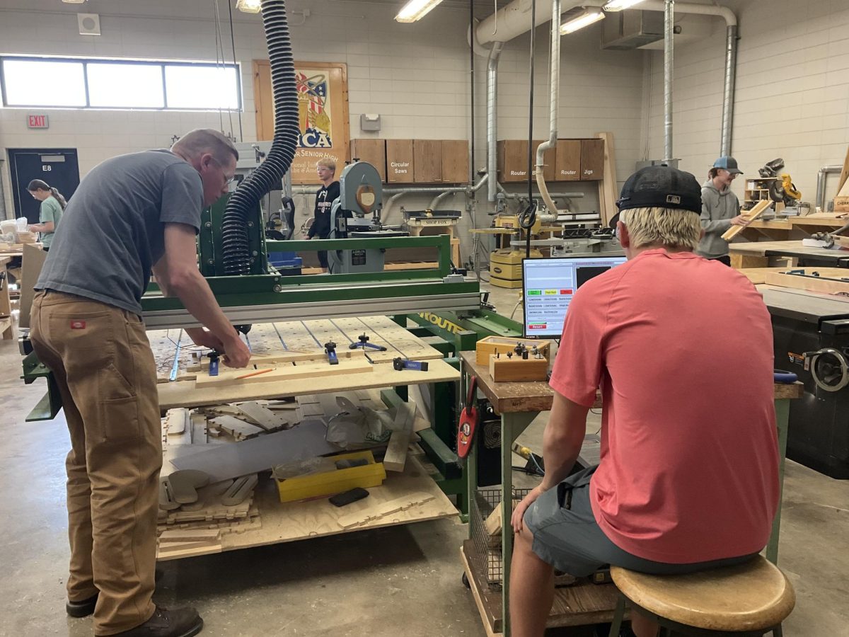  Woods teacher Adam Kuhlman (right) shows freshman Luke Lundgren (left) how to use the CNC router in the woodshop on May 12, 2025. Kuhlman helps the younger students learn and grow in class. 