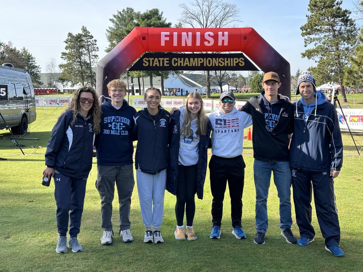 Smith (second on the right) poses in front of the cross country State banner with four current athletes and head coach Lee Sims in Wisconsin Rapids on Nov. 2. 