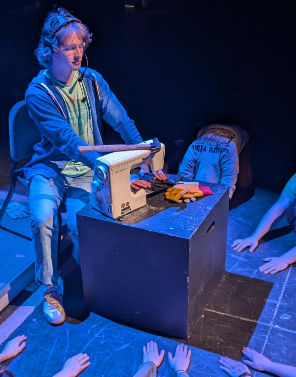 Senior Wyld Wittkopf sews up a costume on stage with a hammer and a pair of lighting gloves resting on a table, April 14, 2025. This is Wittkopf’s first time as Costume manager for a performance.