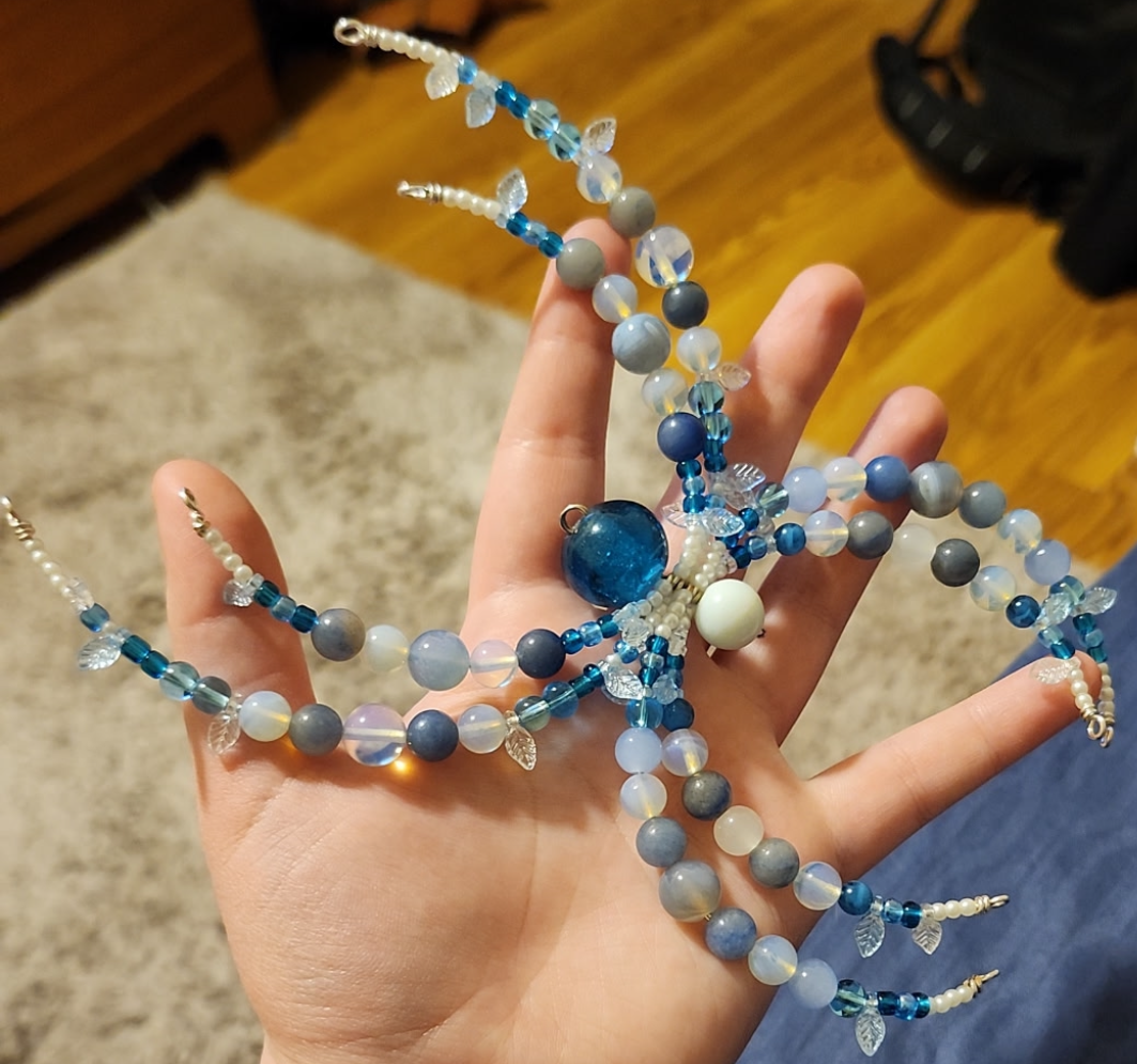 A person holds up an ornate, handcrafted spider ornament. The spider is made of several dozen colored beads.