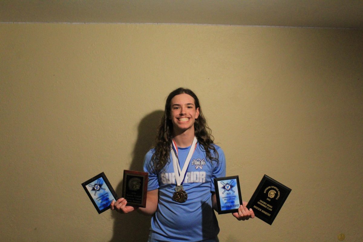 Senior Breckin Bergquist stands in his room with two football plaques, two baseball plaques, three other medals, and wearing his Strike Out Cancer baseball jersey. Showing athletic success that he has made during high school. April 13, 2026.