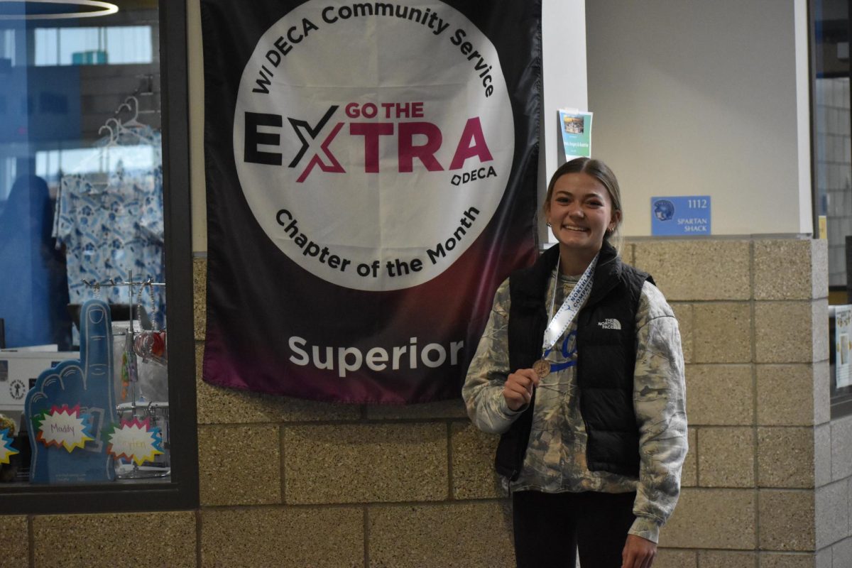 Freshman Anna Staupe poses in front of the DECA Chapter of the month sign, holding up her figure skating medal on Mar. 30.